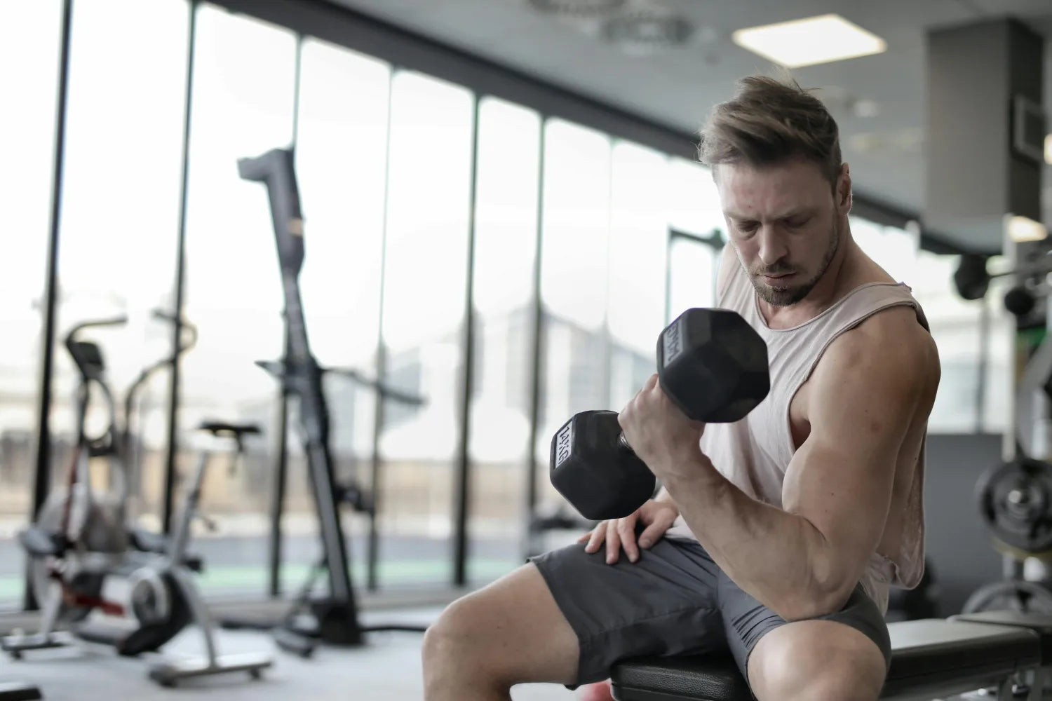 Homme musclé s'entraînant en salle de sport avec un haltère, effectuant un curl biceps sur un banc, dans un environnement lumineux et moderne.